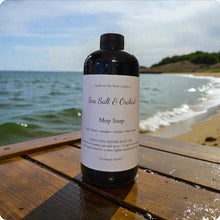 Load image into Gallery viewer, Mop soap bottle labeled 'Sea Salt & Orchid' on a wooden surface with a beach background