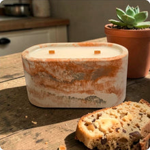 Load image into Gallery viewer, Candle in a rustic concrete container on a wooden surface with banana nut bread and a potted plant in the background
