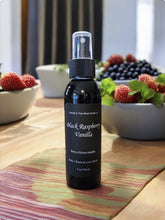 Load image into Gallery viewer, A black spray bottle labeled 'Black Raspberry Vanilla' positioned on a table with a bowl of berries, a bottle of wine, and raspberries in the background.
