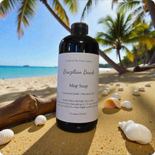 Load image into Gallery viewer, Bottle of Brazilian Beach Mop Soap on a sandy beach with palm trees and shells.
