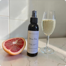 Load image into Gallery viewer, Spray bottle labeled 'Citrus Kiss' next to a half-cut grapefruit and a glass of sparkling wine on a marble surface.
