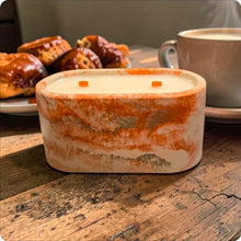 Load image into Gallery viewer, Concrete candle with marbled design on a wooden surface with pastries and a mug in the background.
