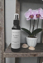 Load image into Gallery viewer, Bottle of 'Sea Salt &amp; Orchid' body spray on a wooden shelf with an orchid plant and shells.
